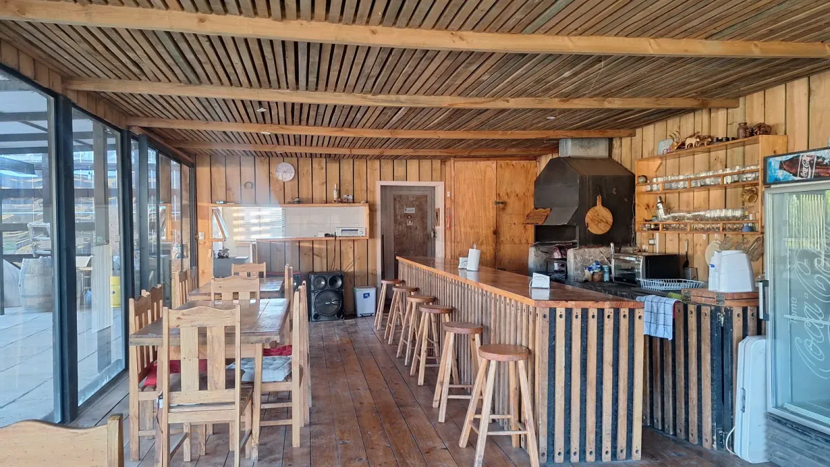 Interior del quincho con mesas de madera, barra y ventanales al campo